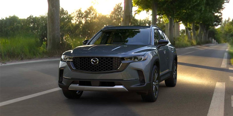 Gray Mazda CX-70 SUV driving along a tree-lined road in daylight