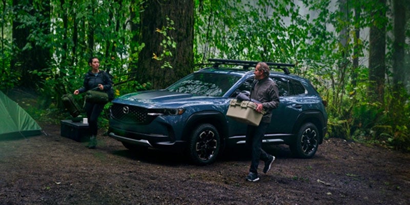 Mazda CX-70 SUV parked at a forest campsite with outdoor gear, as campers unload equipment among tall trees