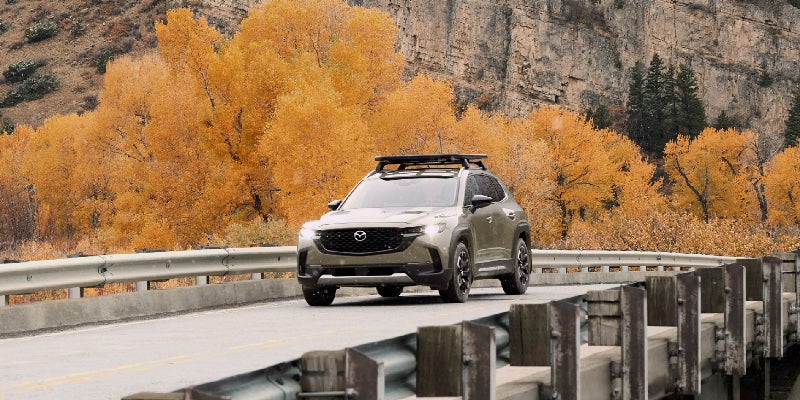 Mazda CX-70 SUV equipped with a roof rack driving across a bridge, surrounded by autumn-colored trees