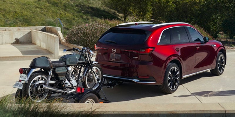 Red Mazda CX-90 SUV towing a motorcycle on a trailer along a paved road in a scenic outdoor setting