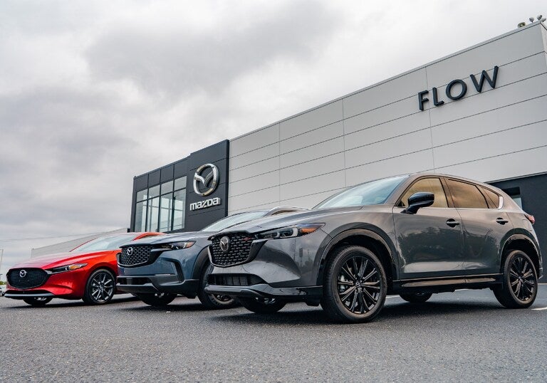 Gray Mazda CX-5 SUV and red Mazda sedan parked outside a Mazda dealership showroom