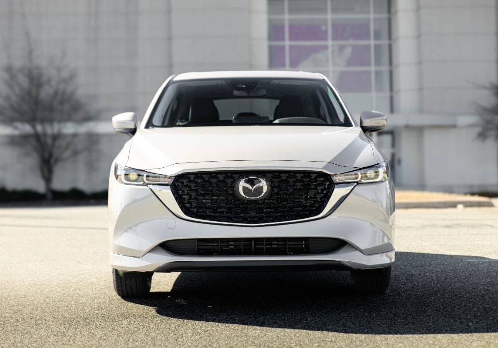White 2025 Mazda Cx-5 SUV parked in a lot with a large building in the background