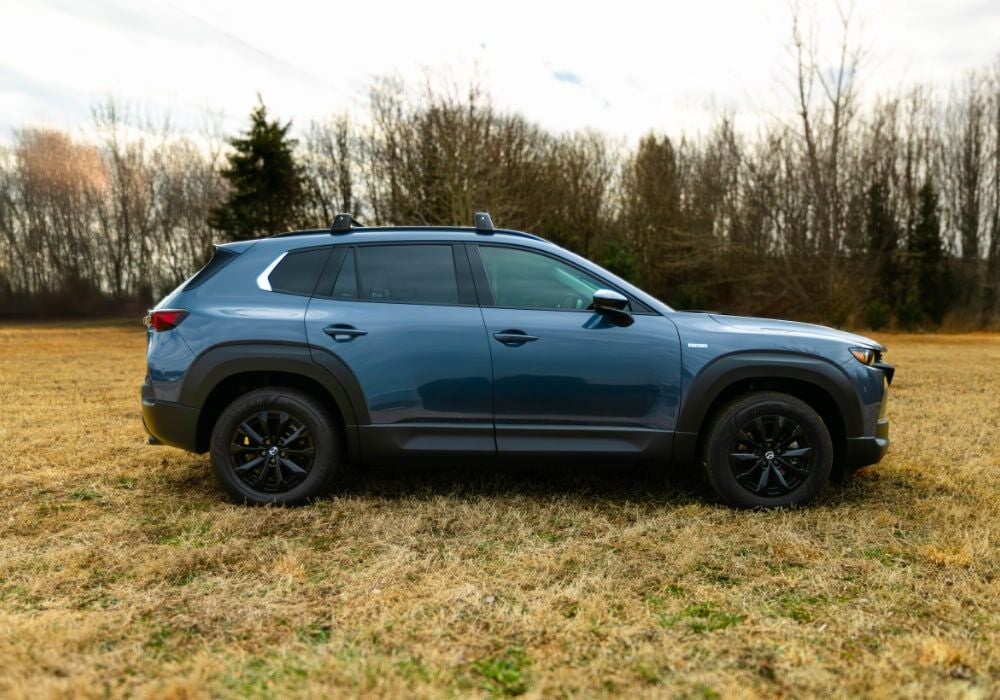 Passenger side view of a 2025 Mazda CX-50 parked in a grassy field with trees in the background