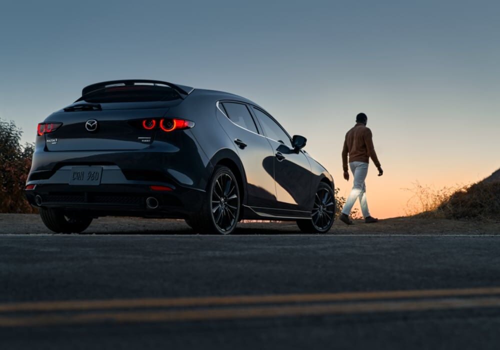 Mazda3 Hatchback parked on the side of the road facing away from the camera. There is a person walking