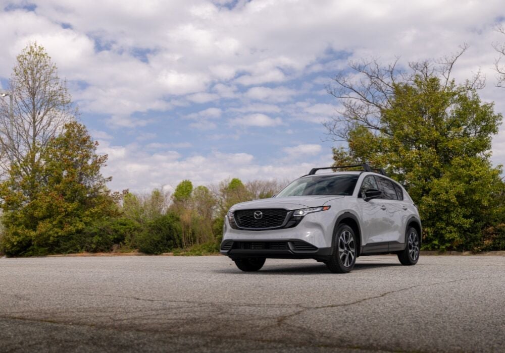 Front view of a 2026 Mazda CX-5 SUV parked in an empty lot. The SUV is light gray and there are tree…